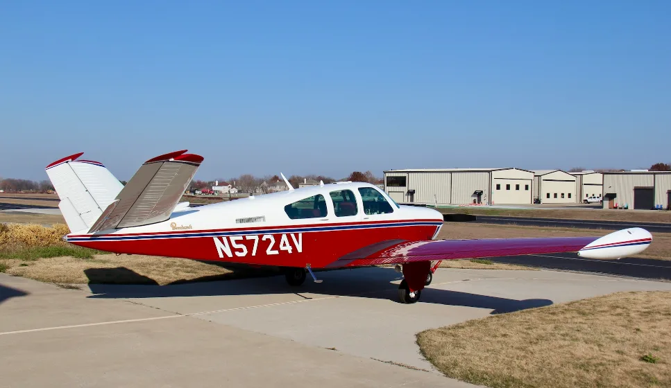 A red and white Beechcraft Bonanza N5724V parked on an airfield with hangars in the background.