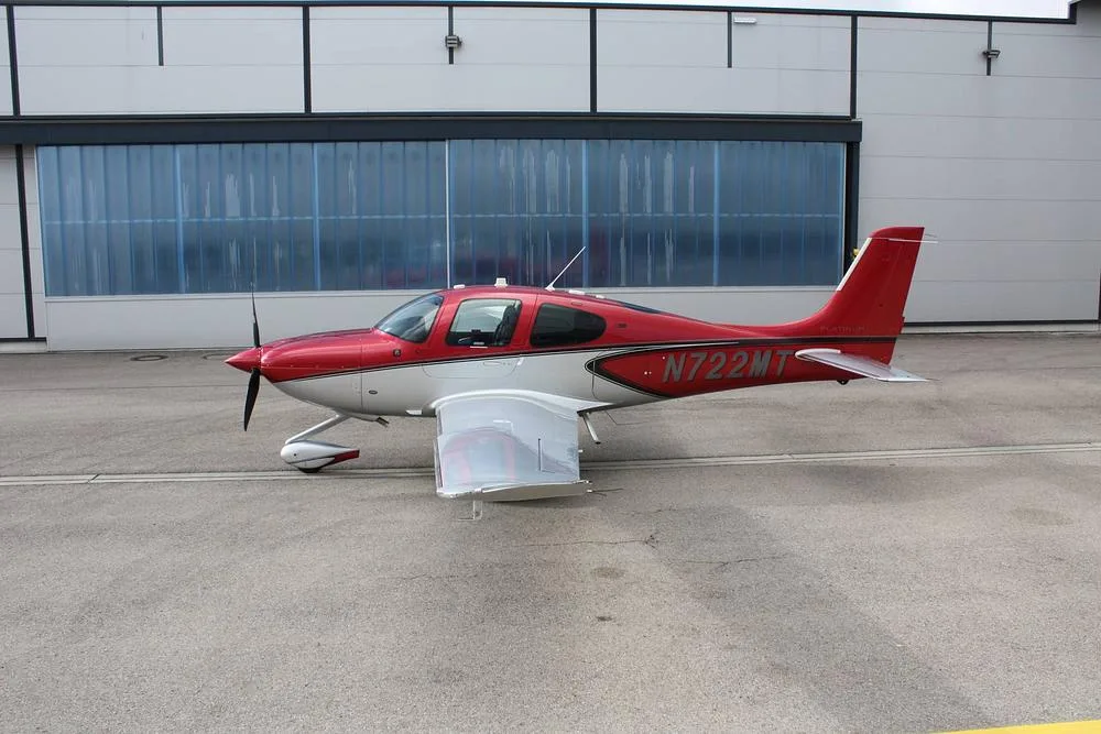 A red and white Cirrus SR22 aircraft, registration N722MT, parked on an asphalt apron in front of a hangar.