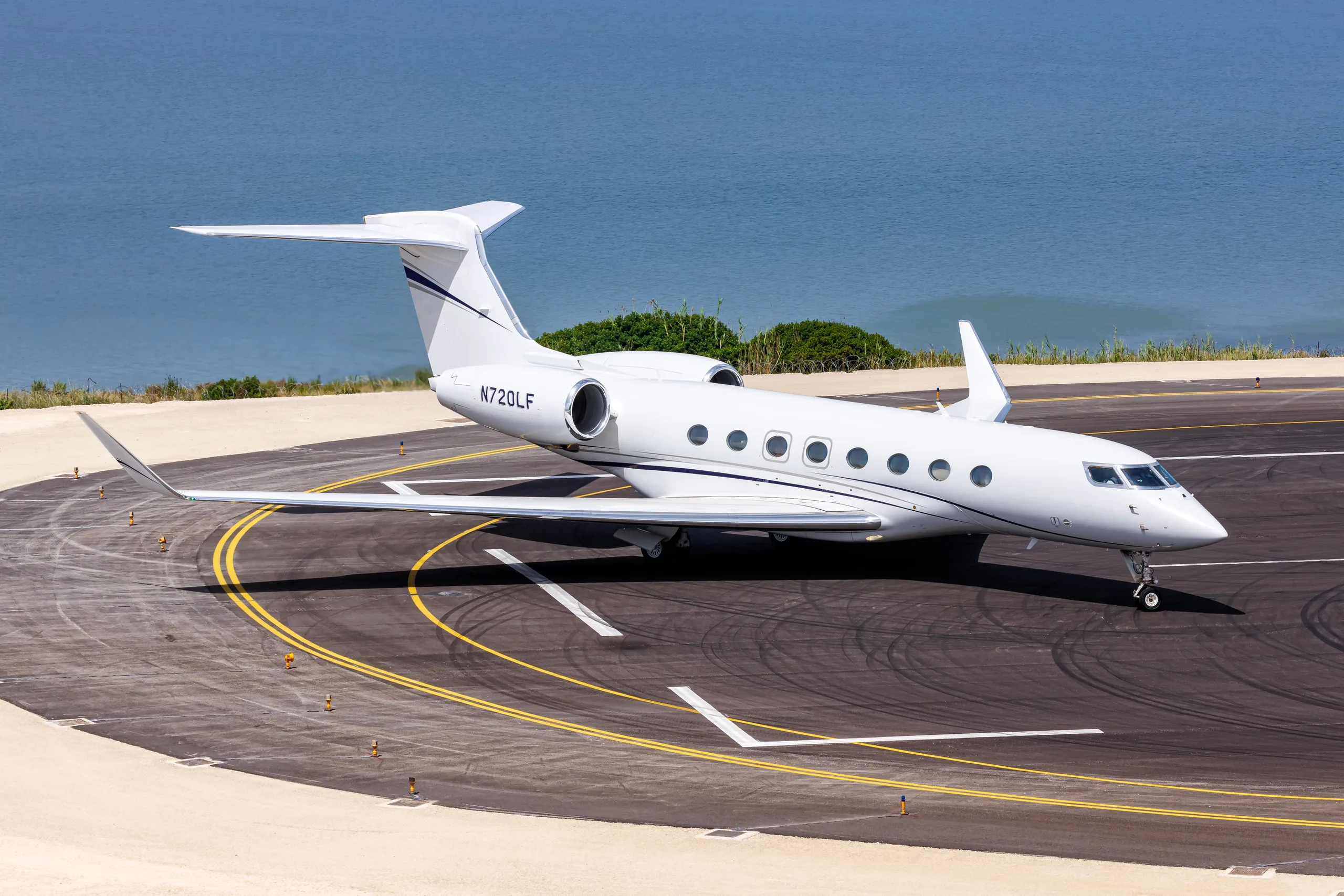 A white Gulfstream G650 business jet with blue accent stripes is parked on an airfield with the ocean in the background.