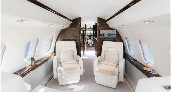 A view of the luxurious white interior cabin of a private jet, featuring cream-colored seats and a screen in the background.