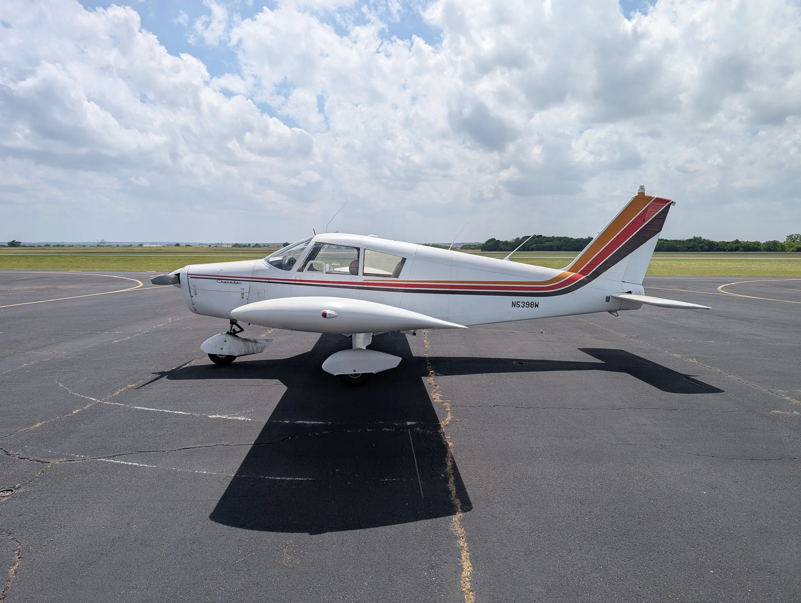 A white Piper Cherokee aircraft with brown and orange stripes on the tail and fuselage, parked on an asphalt runway under a partly cloudy sky.