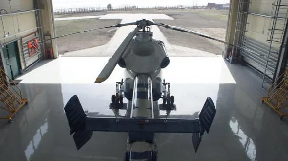 A silver helicopter, viewed from the rear, inside a hangar with the doors open to an airfield.