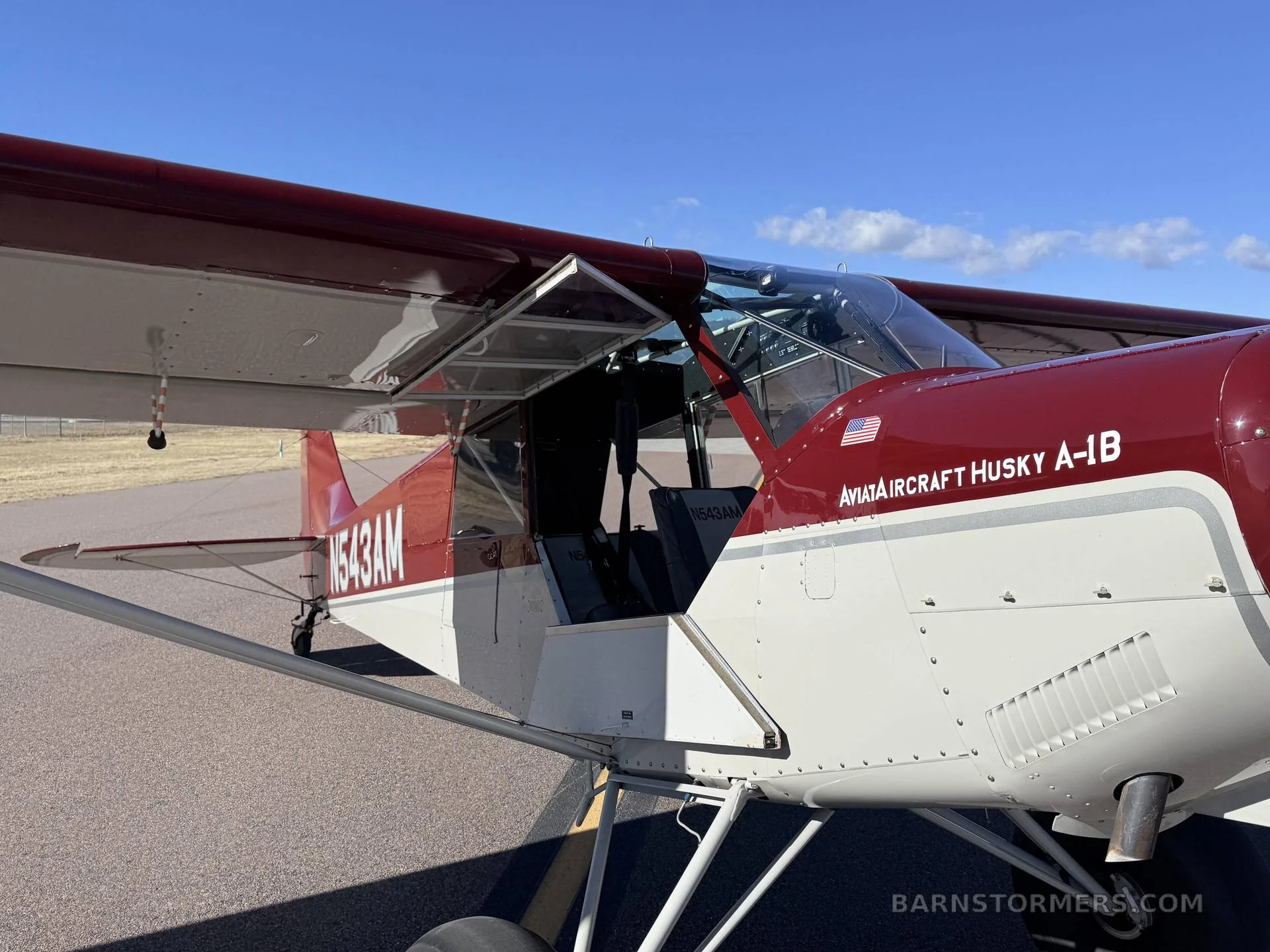 A close-up view of the red and white Aviat Husky A-1B aircraft, showing the open door and cockpit.