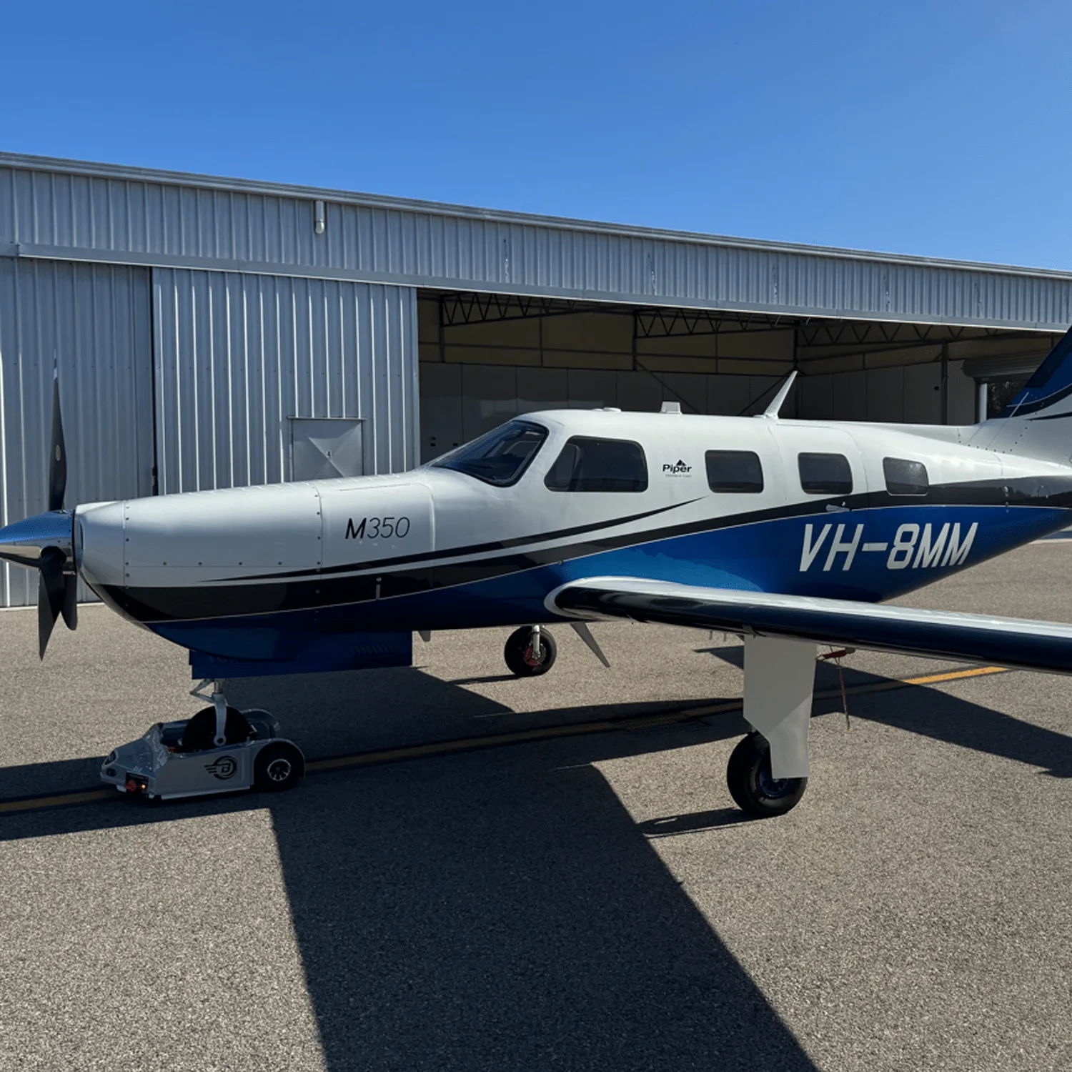 A white and blue Piper M350 aircraft with registration VH-8MM parked on an airfield in front of a hangar.