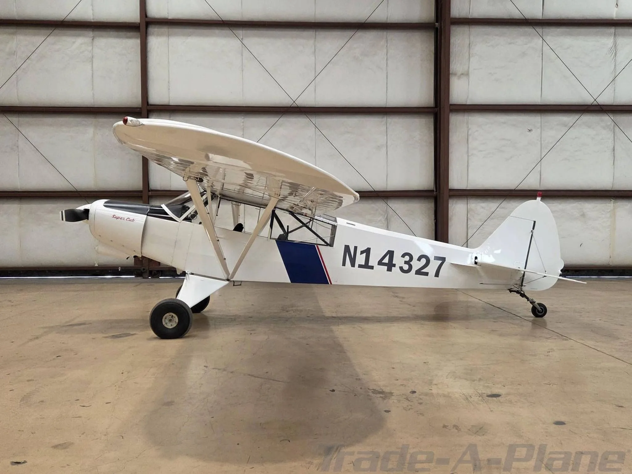 A white Piper Super Cub aircraft with blue and red stripes, registration N14327, parked in a hangar.
