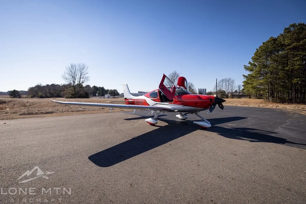 A red and silver Cirrus SR22T aircraft with open doors parked on an asphalt ramp with trees in the background.
