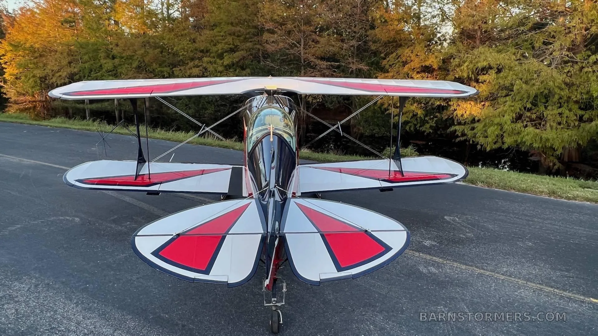 A dark blue, red, and white biplane is parked on an asphalt surface, viewed from the rear with autumn trees in the background.