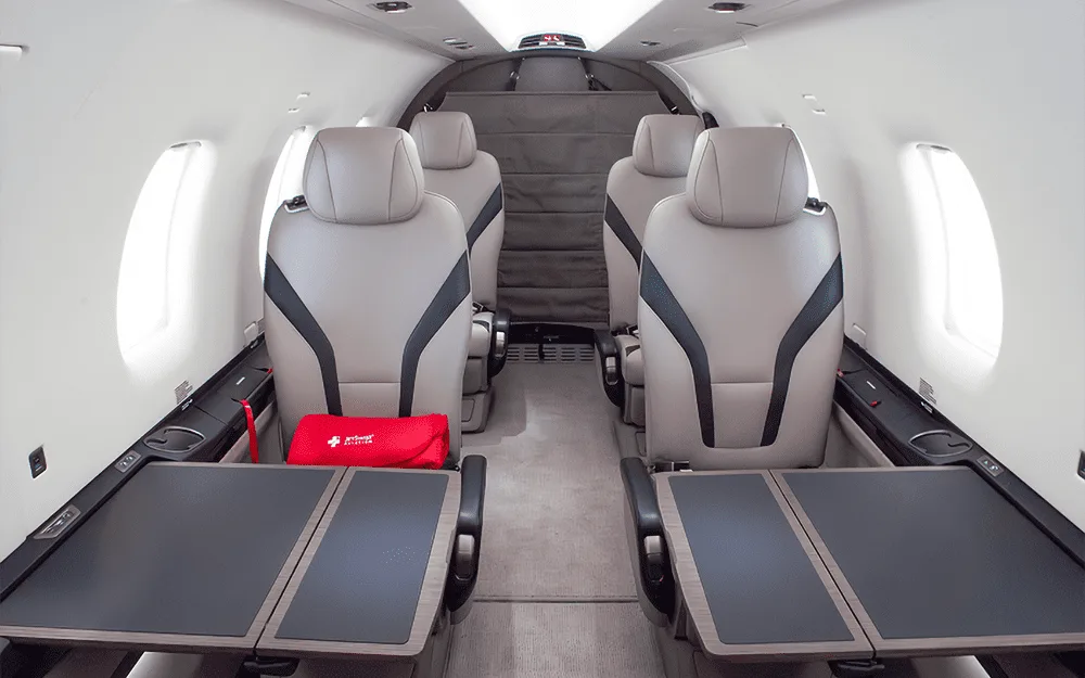 The interior cabin of a Pilatus PC-12 aircraft showing light grey seats with black accents and fold-out tables.