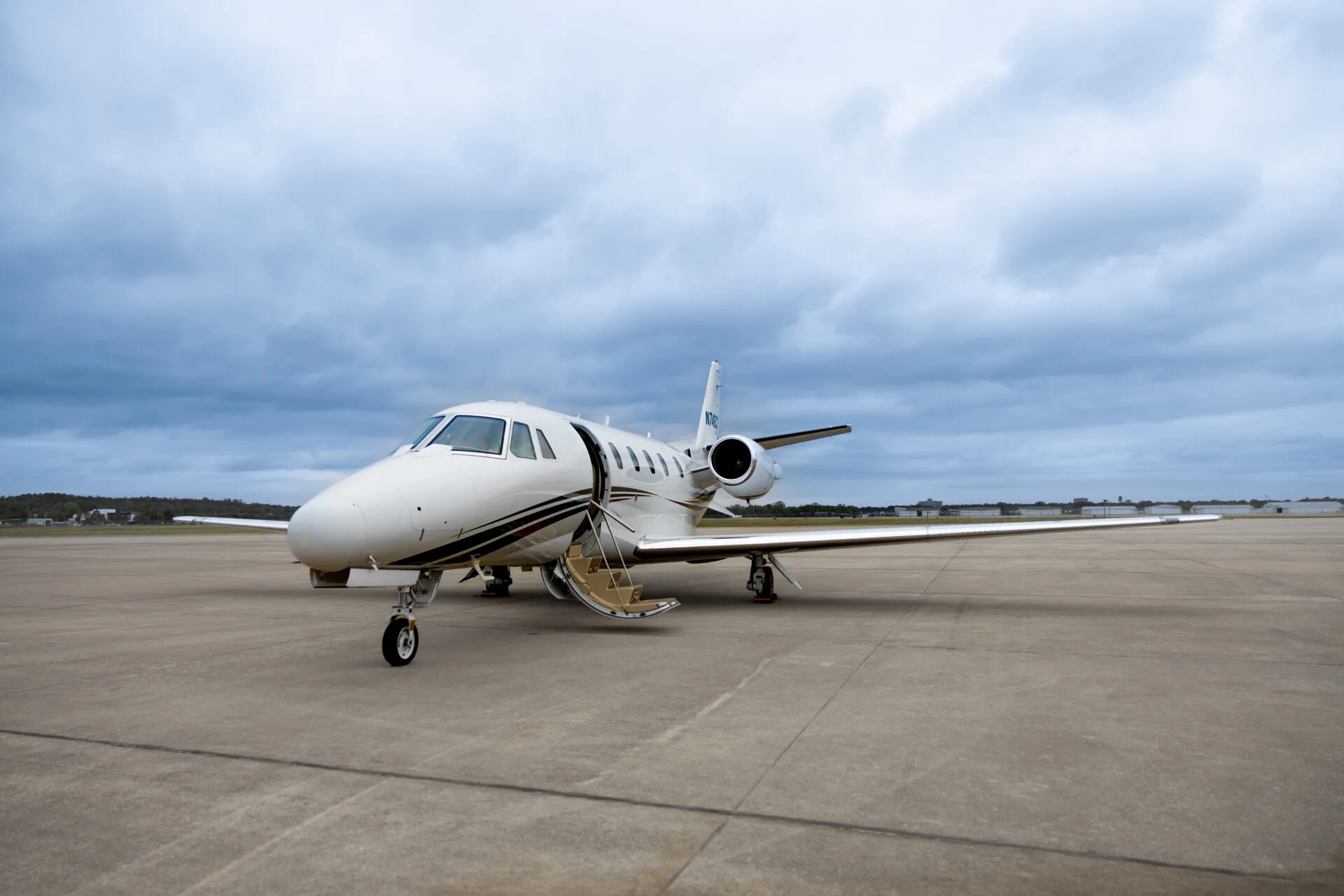 A white business jet with black and gold stripes, tail number N707X, parked on an airfield with its door open under a cloudy sky.