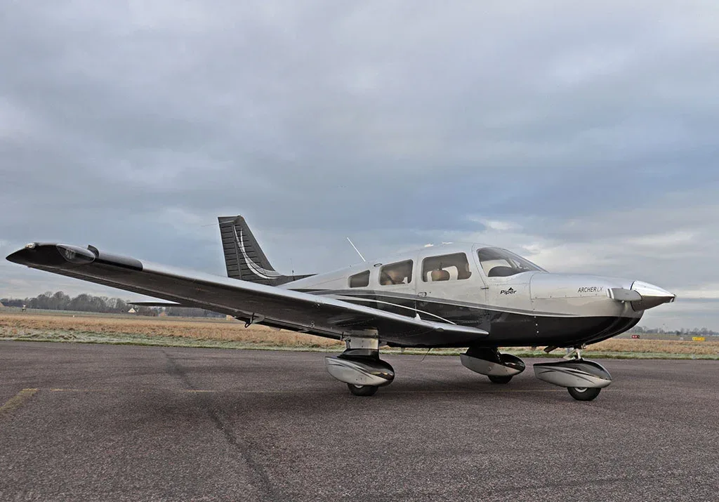 A side view of a silver and black Piper Archer LX aircraft on an airfield under an overcast sky.