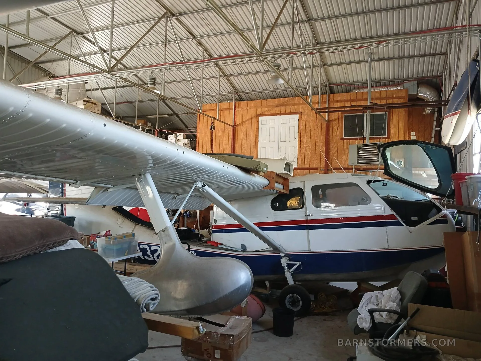 A white, red, and blue amphibious aircraft with floats, partially disassembled, inside a hangar.