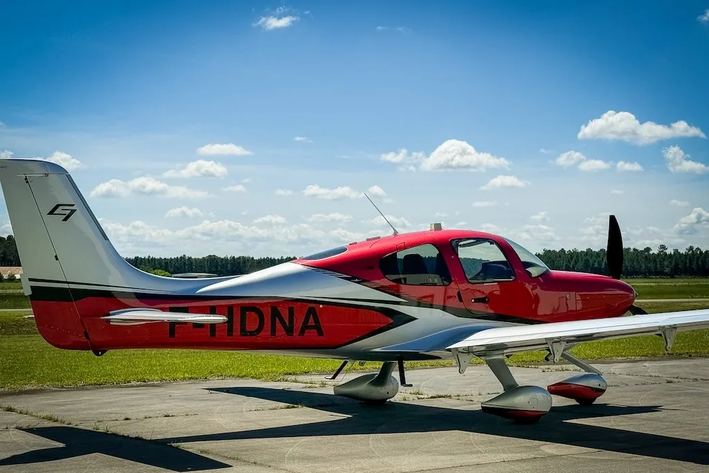 A red and silver Cirrus SR22 GTS (F-HDNA) aircraft parked on a tarmac, viewed from the side.