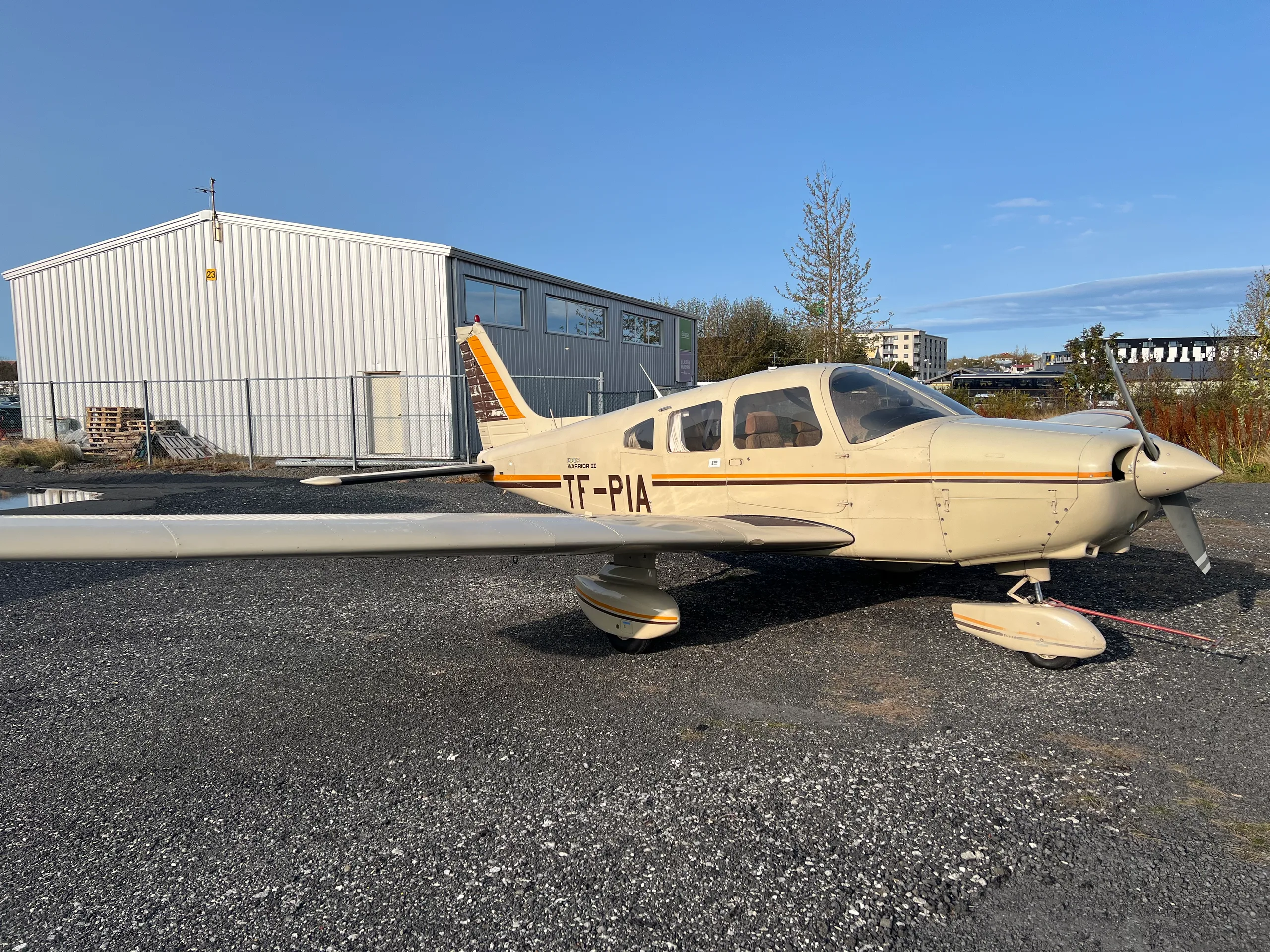A beige Piper Warrior II aircraft with orange and brown stripes is parked on a gravel lot next to a hangar under a clear blue sky.