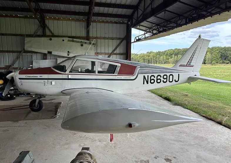 A three-quarter side view of the white and maroon Piper Cherokee 140, N6690J, parked in a hangar.