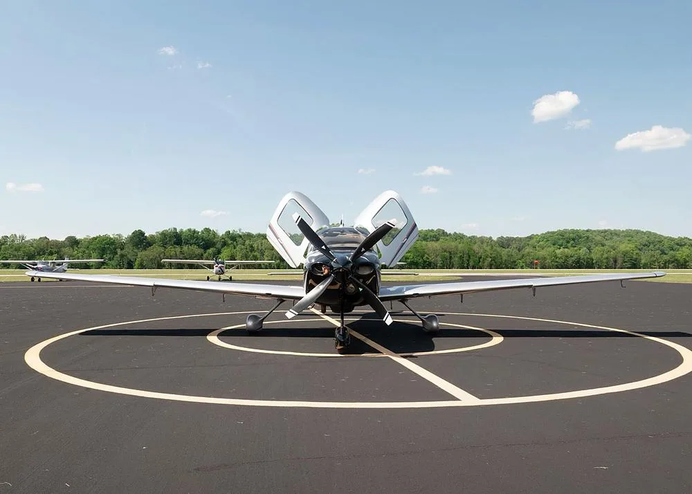 A front view of the silver and black Cirrus SR22 aircraft N816RJ parked on an airfield with its gull-wing doors open, other aircraft in the background.