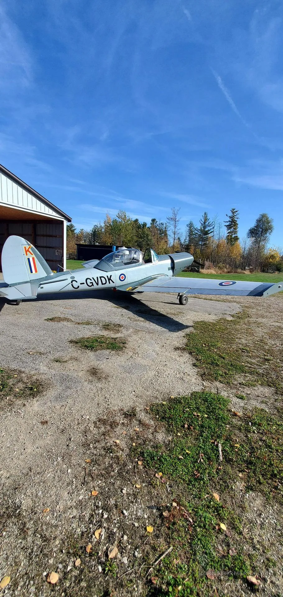 A light grey single-engine aircraft with Canadian registration C-GVDK parked on a gravel area next to a hangar under a clear blue sky.