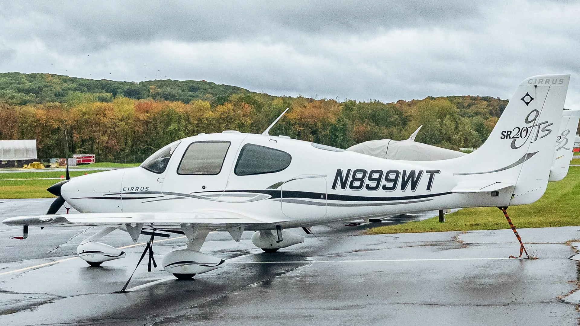 A white Cirrus SR20 aircraft with black and grey accents, registration N899WT, parked on a wet tarmac with autumn trees in the background.