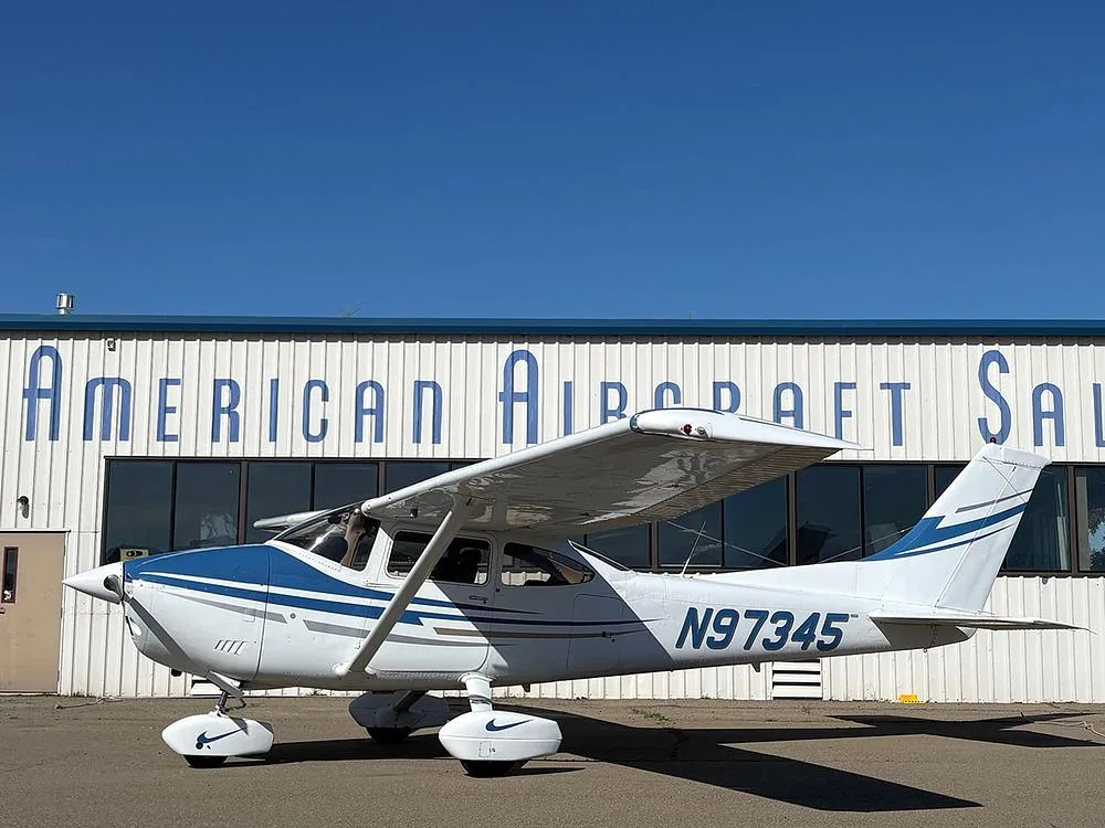 A white and blue Cessna aircraft with registration N97345 parked on an tarmac in front of the American Aircraft Sales building.