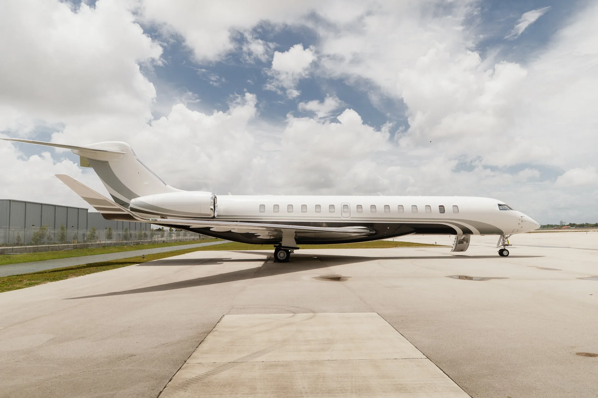 A white business jet with grey and black accents is parked on a tarmac under a partly cloudy sky.