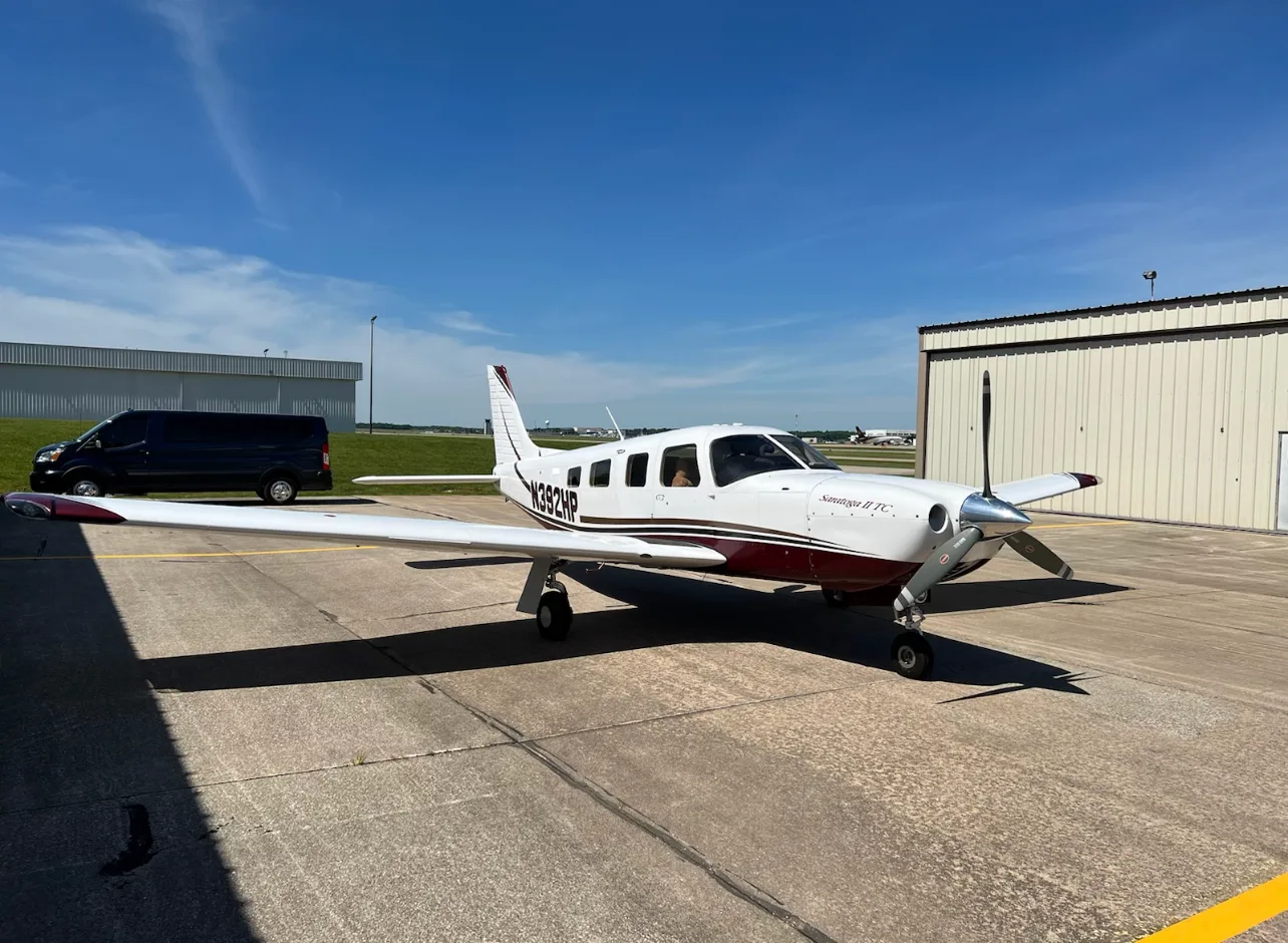 A white and maroon Piper Saratoga II TC is parked on an airport tarmac on a sunny day.