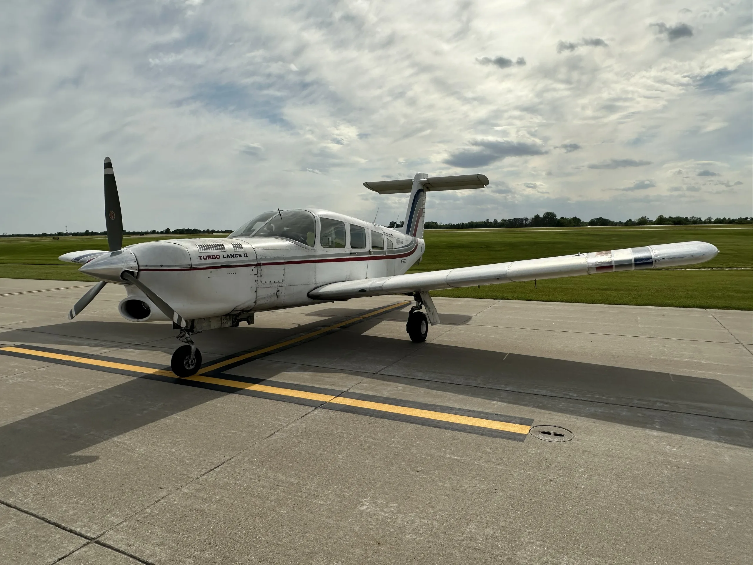 A white Piper Turbo Lance II aircraft with red and blue stripes parked on an airfield, showing the right side.