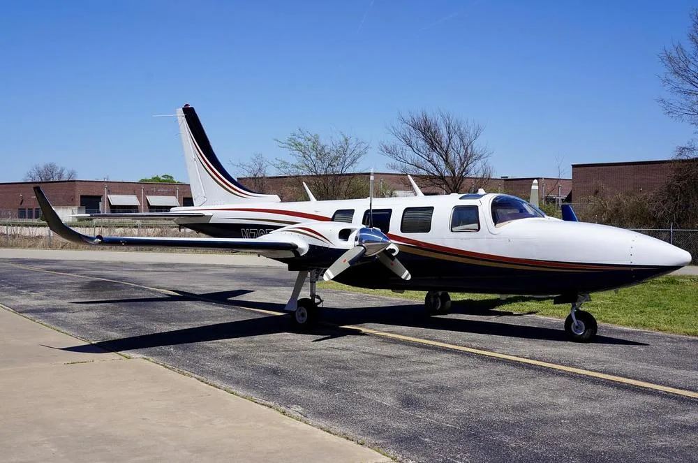 A white and dark blue twin-engine propeller aircraft with gold and red stripes, parked on an asphalt ramp under a clear blue sky.