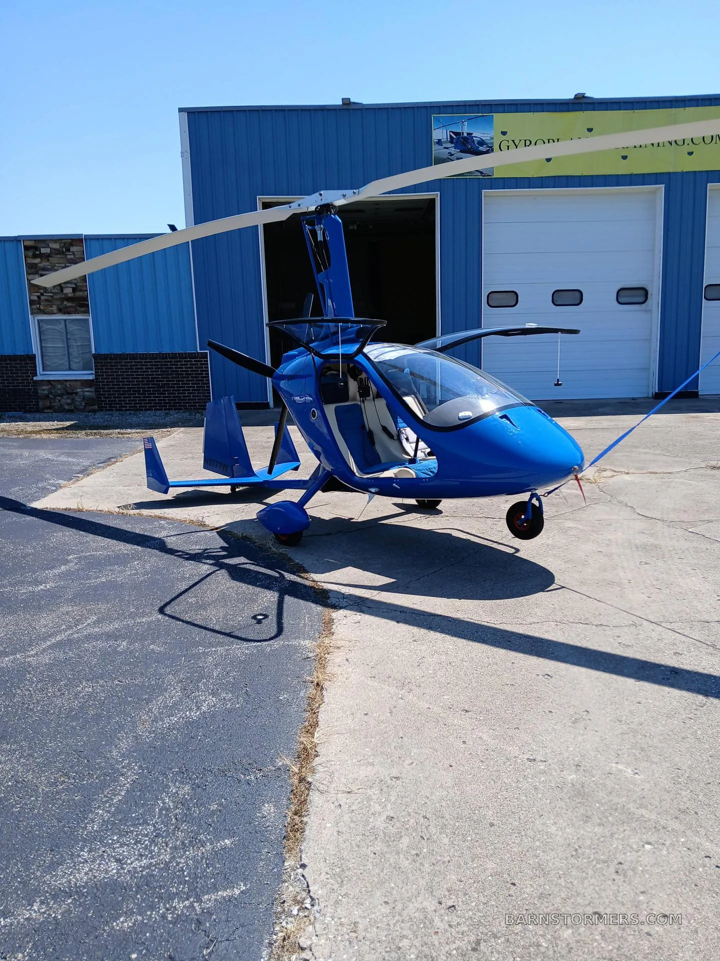 A blue gyrocopter with an open canopy parked on a tarmac in front of a blue hangar.