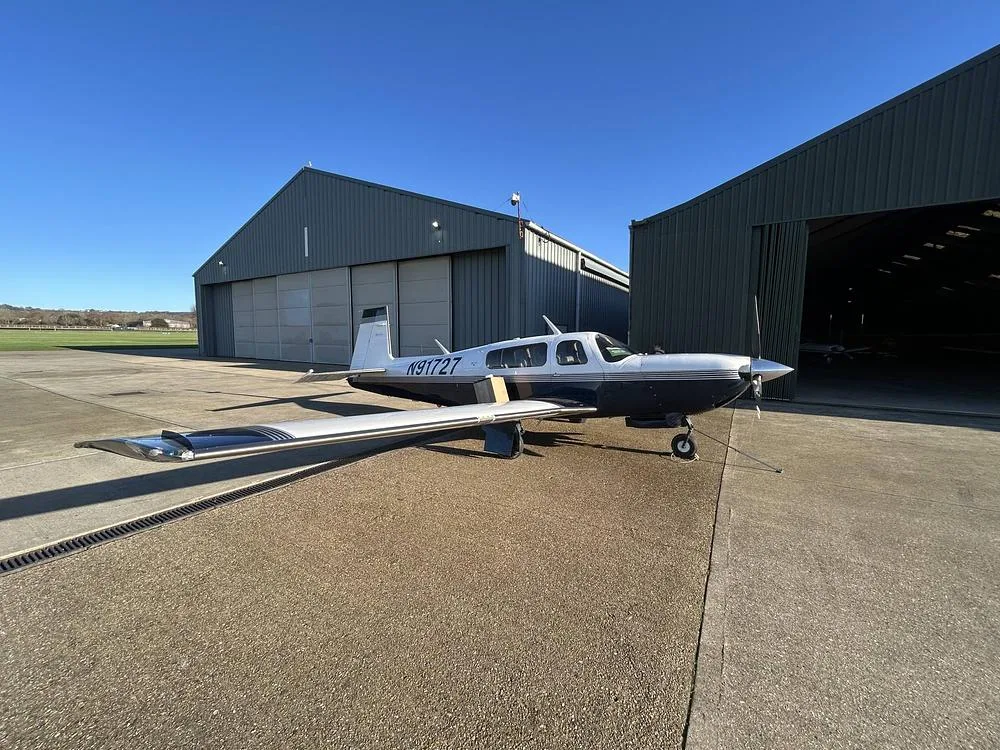 A white and blue single-engine aircraft, N91727, is parked on an asphalt ramp in front of two hangars under a clear blue sky.