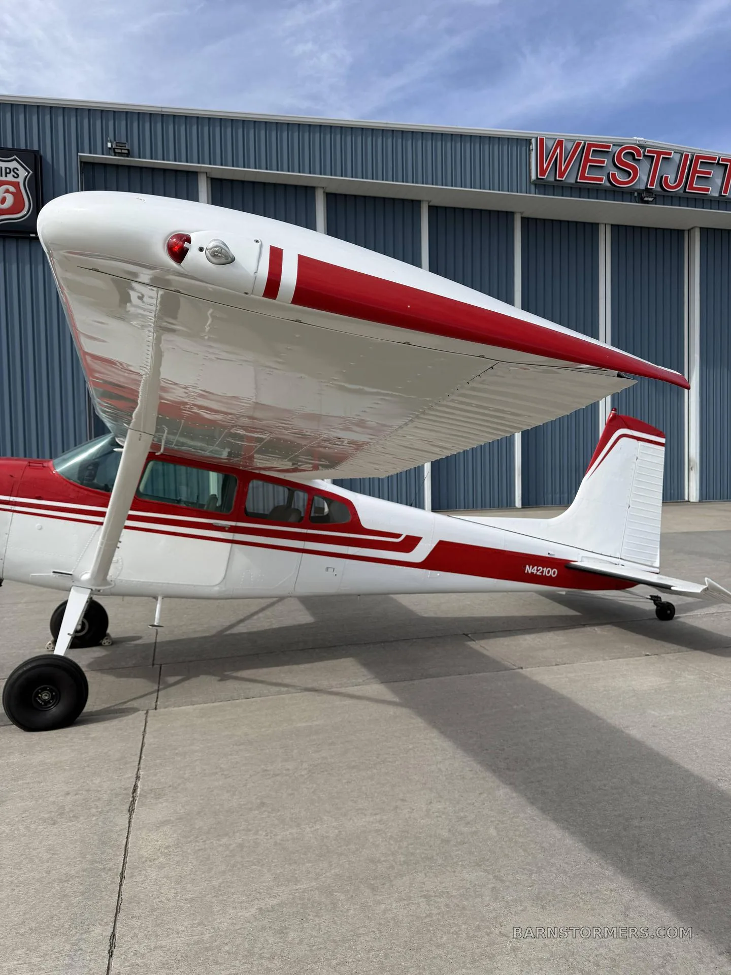 A side view of a white and red single-engine propeller aircraft (N42100) parked in front of a blue Westjet hangar.