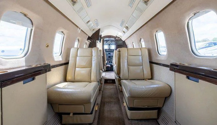 The interior cabin of a business jet, showing rows of beige leather seats leading towards the cockpit.