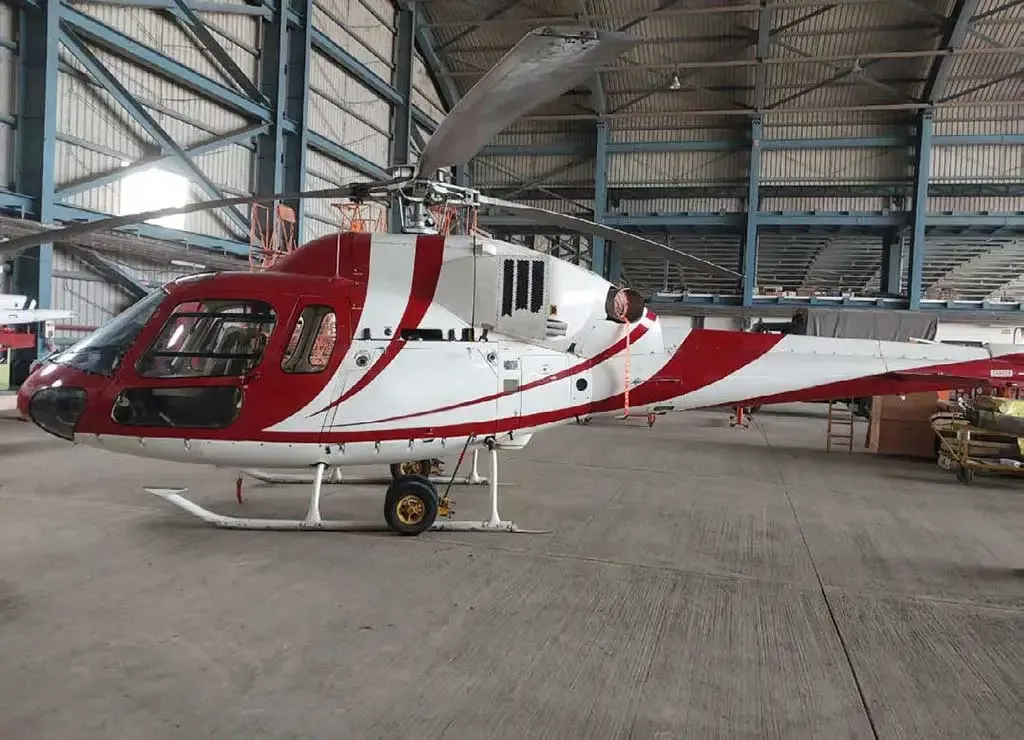 A red and white helicopter with a striped paint scheme parked inside a hangar, viewed from the left side.