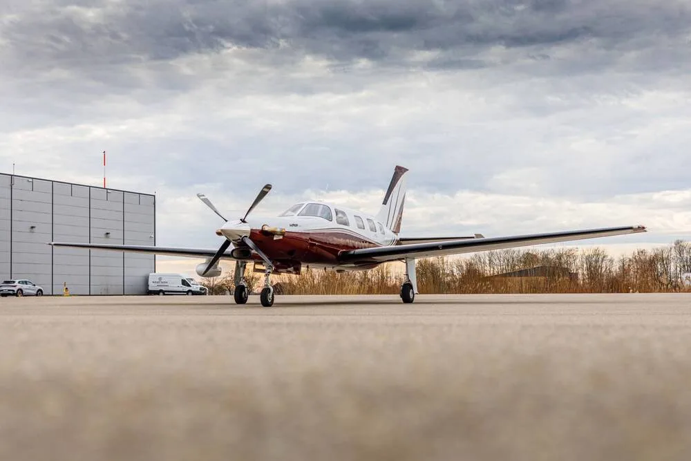 A white and maroon single-engine turboprop aircraft parked on an airfield with a hangar in the background.