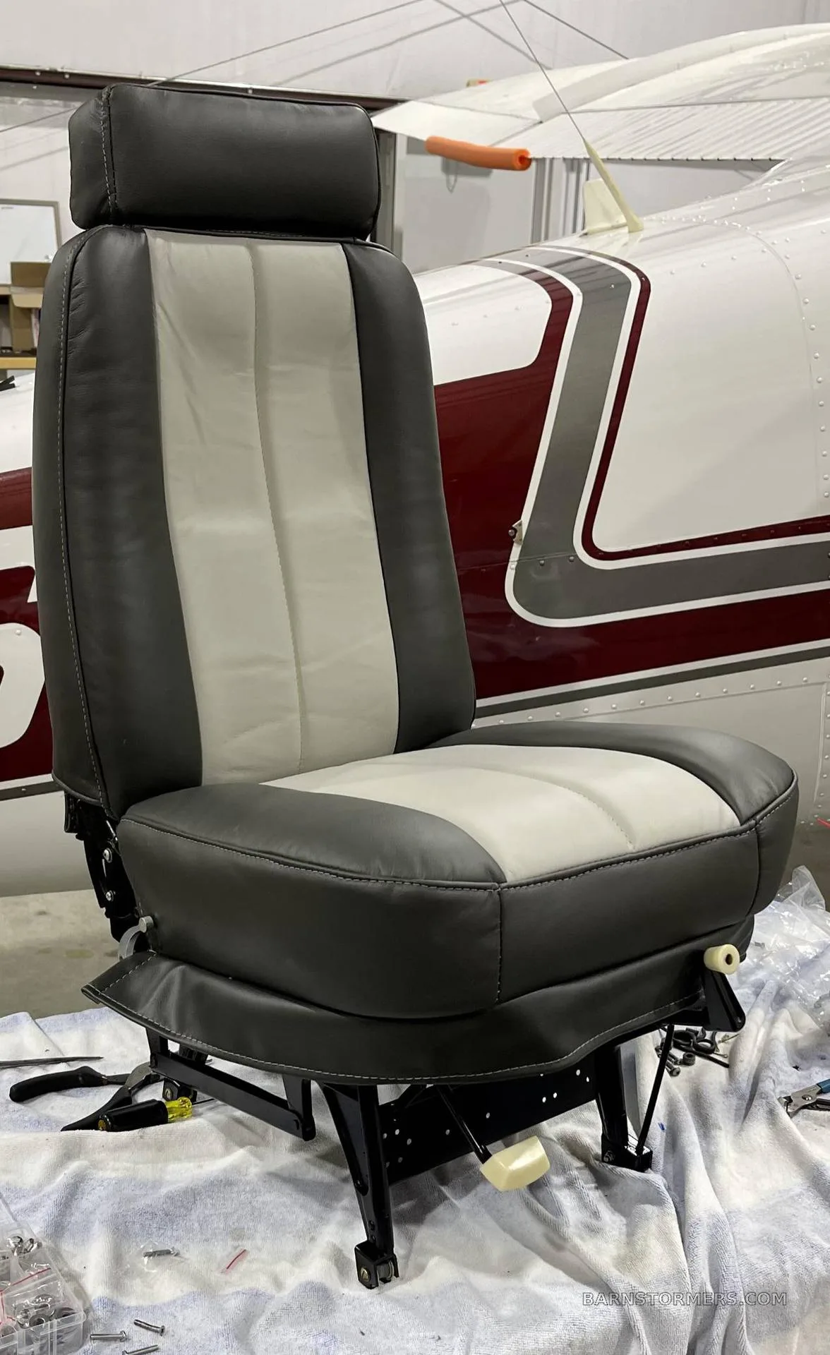 A newly upholstered gray and light gray aircraft seat is shown in front of the fuselage of a white and maroon aircraft.