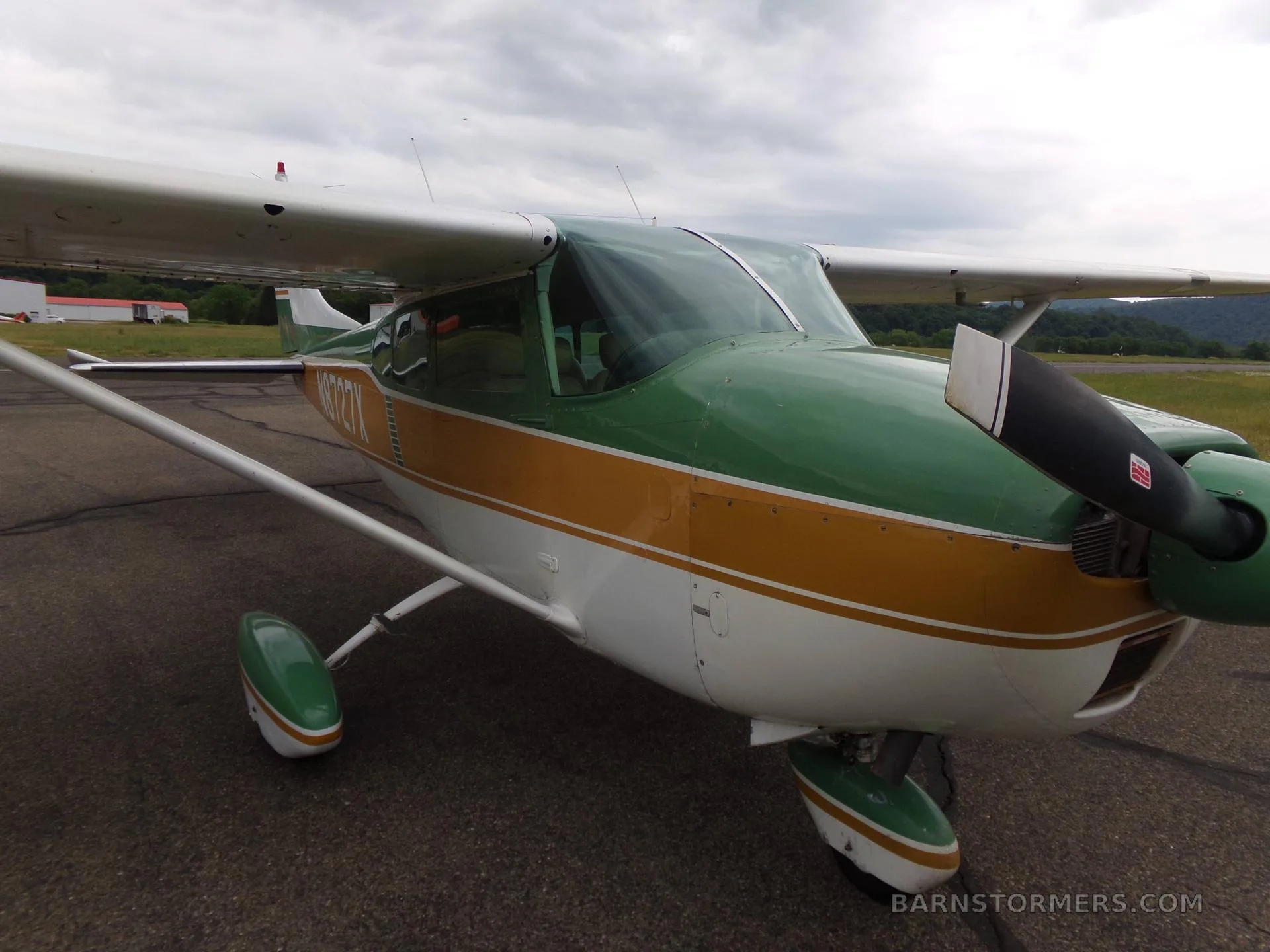 A green, gold, and white Cessna 172 aircraft with registration N8727X, viewed from the front-right on an asphalt ramp under a cloudy sky.