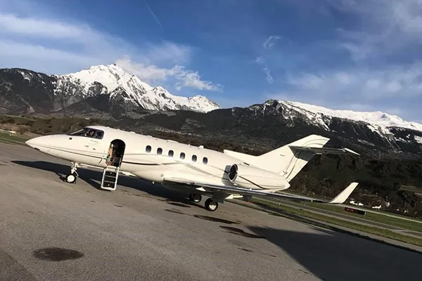 A white private jet with its door open and stairs extended, parked on an tarmac with snow-capped mountains in the background.