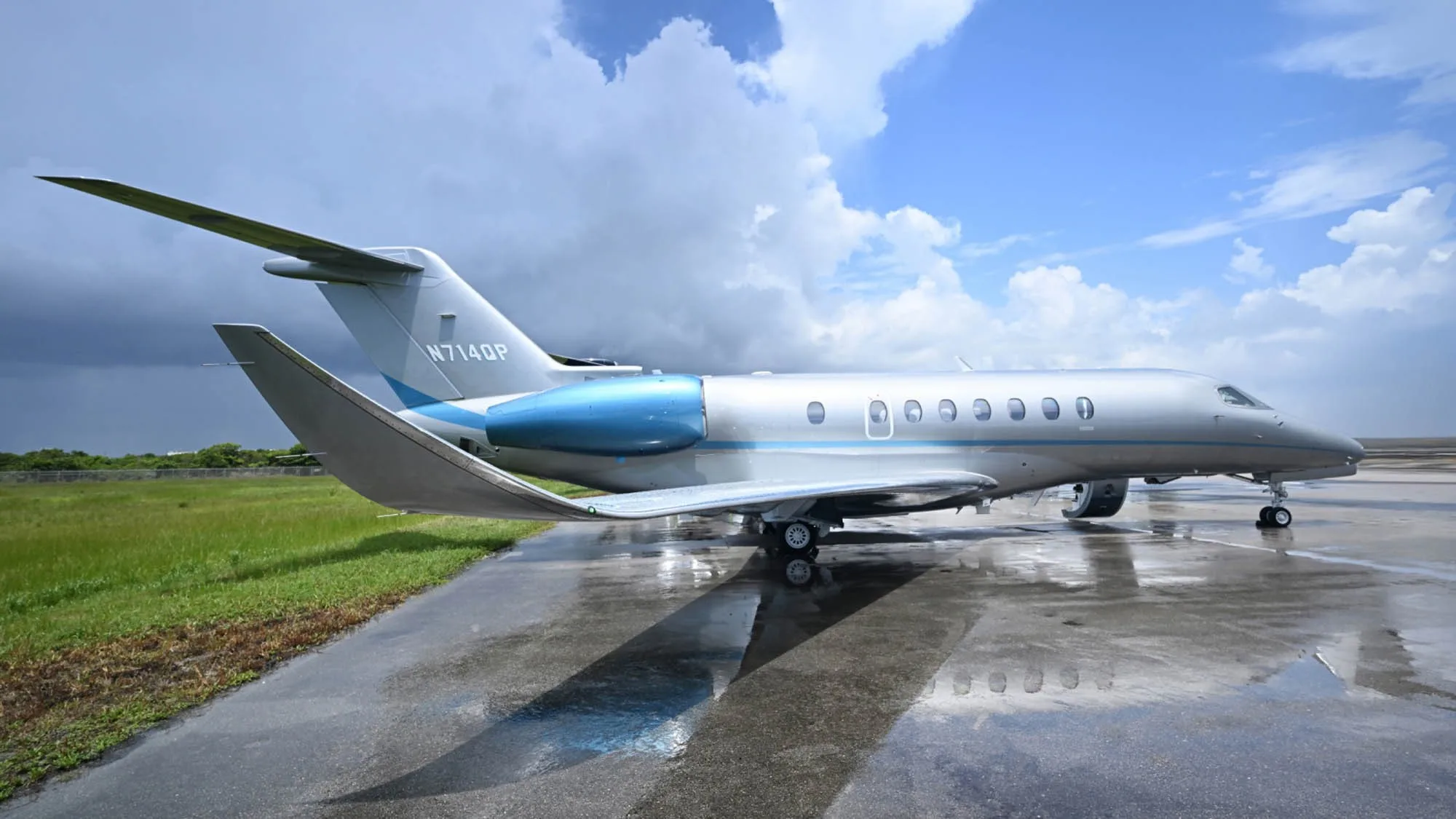 A silver and blue business jet, N714QP, sits on a wet tarmac under a cloudy sky.