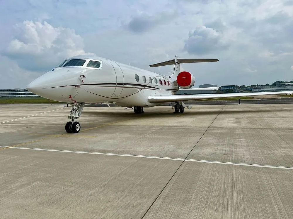 A white Gulfstream G650 business jet with red engine nacelles parked on a concrete tarmac under a cloudy sky.