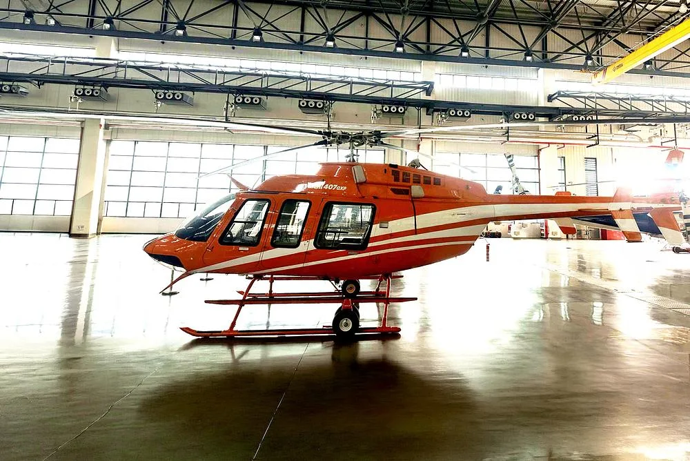 A side view of the orange and white Bell 407GXP helicopter inside a brightly lit hangar.