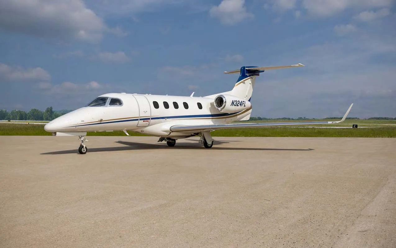 A white Embraer Phenom 300 private jet with blue and gold stripes on the tail and fuselage, parked on an airfield under a partly cloudy sky.