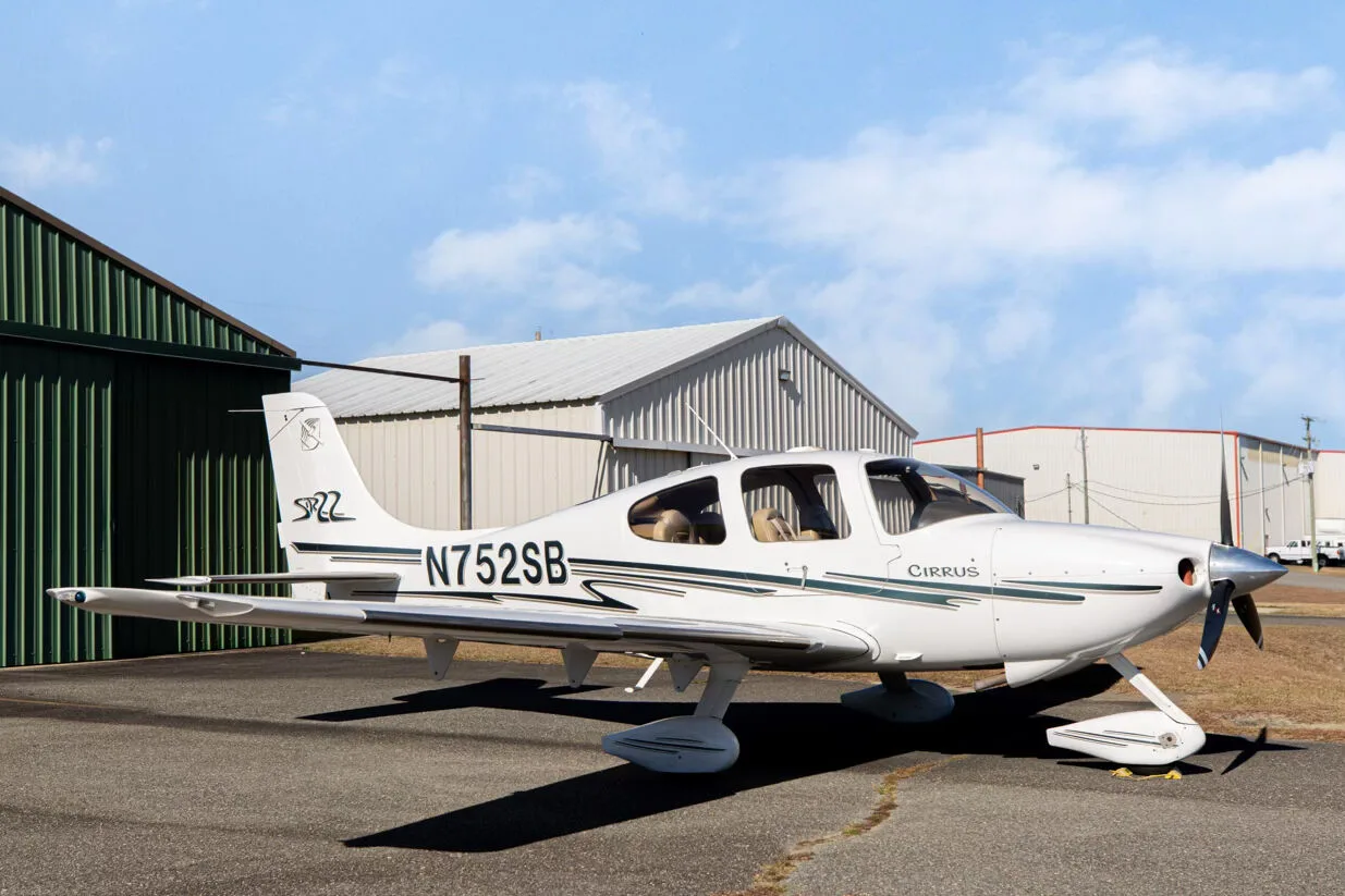 A white Cirrus SR22 aircraft with green and grey stripes, registration N752SB, parked on an asphalt ramp next to a green hangar.