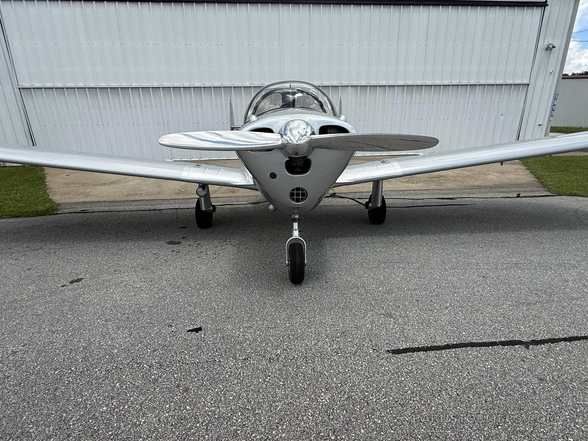 A head-on view of a silver single-engine aircraft with a propeller, parked in front of a white hangar.