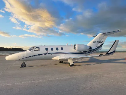 A white Cessna CitationJet with black and blue stripes, tail number N513RV, parked on a tarmac at sunset.