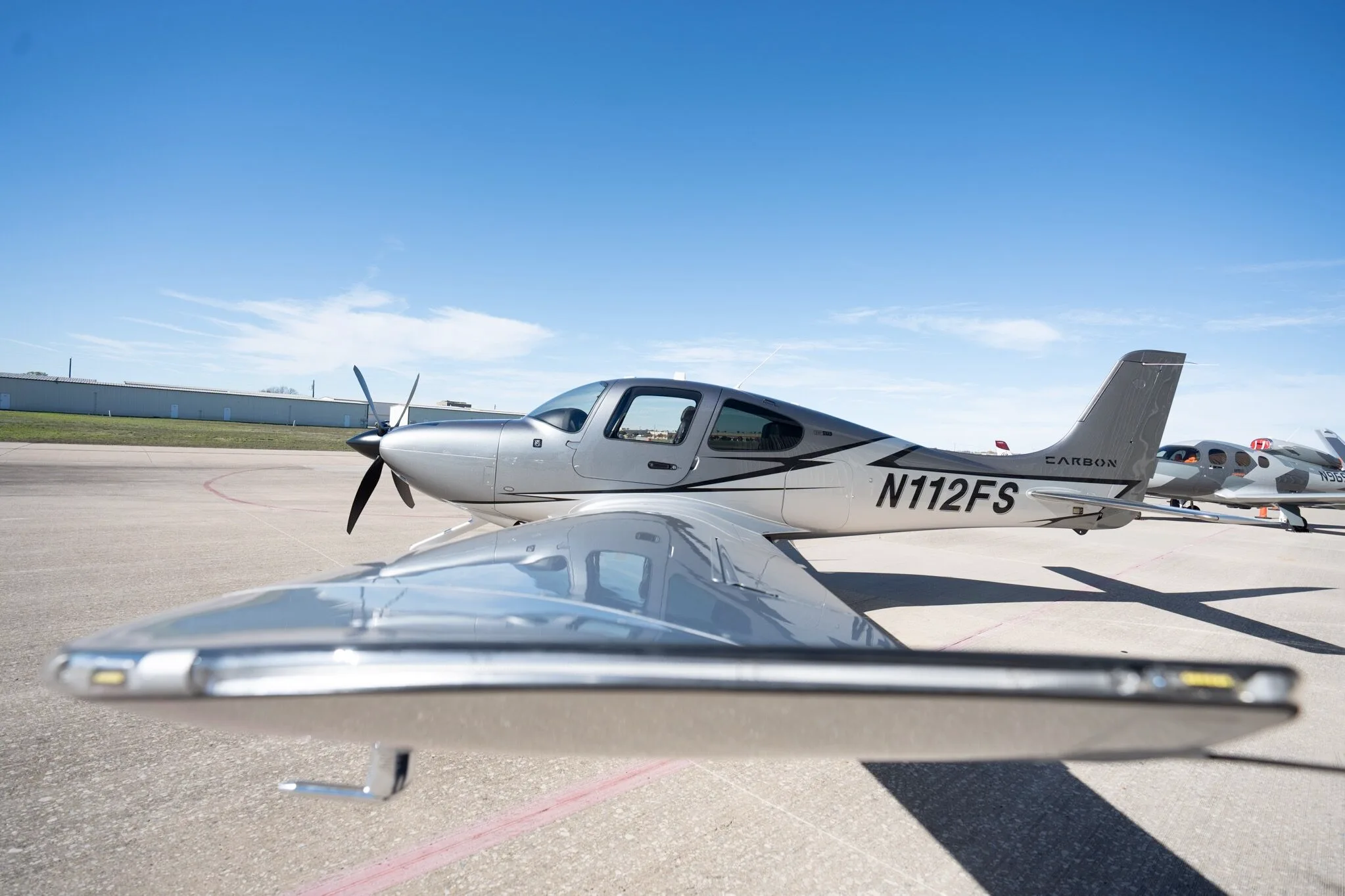A low-angle side view of a silver and dark grey Cirrus SR22T Carbon Edition aircraft (N112FS) on an airport tarmac.