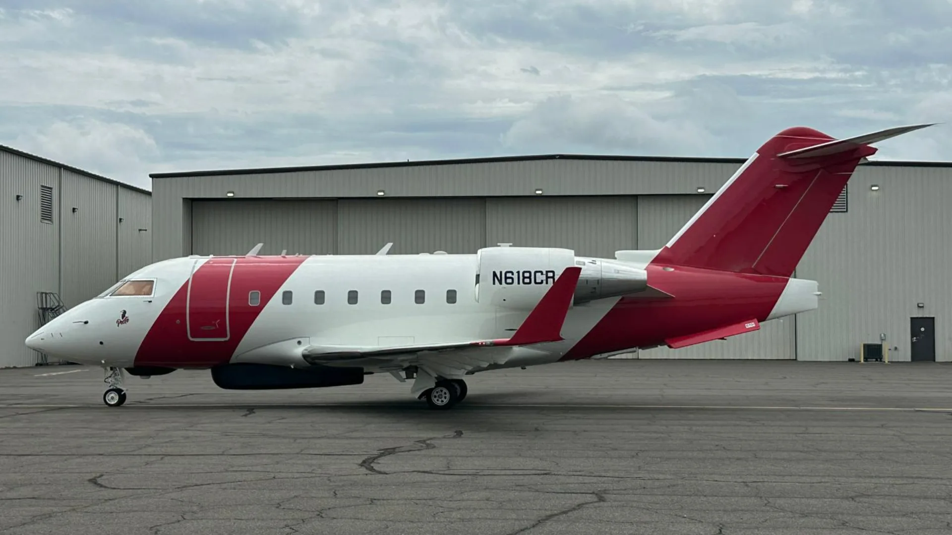 A white and red Bombardier Challenger 604 aircraft, N618CR, parked on an tarmac in front of hangars under a cloudy sky.