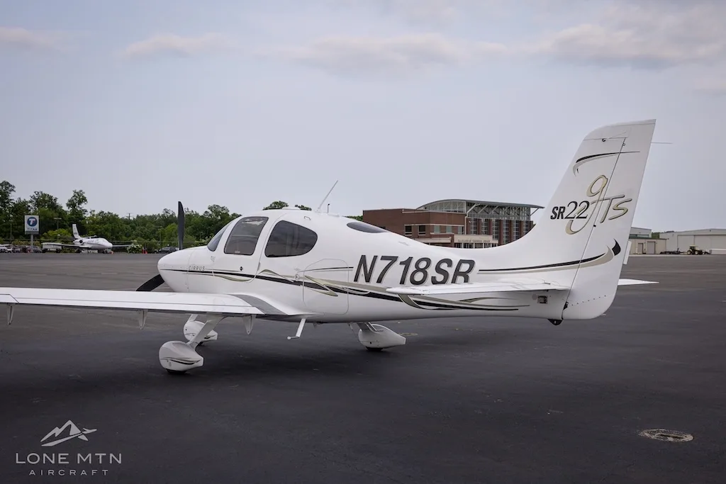 Rear-side view of a white Cirrus SR22T aircraft with black and gold accents parked on an asphalt tarmac with buildings in the background.