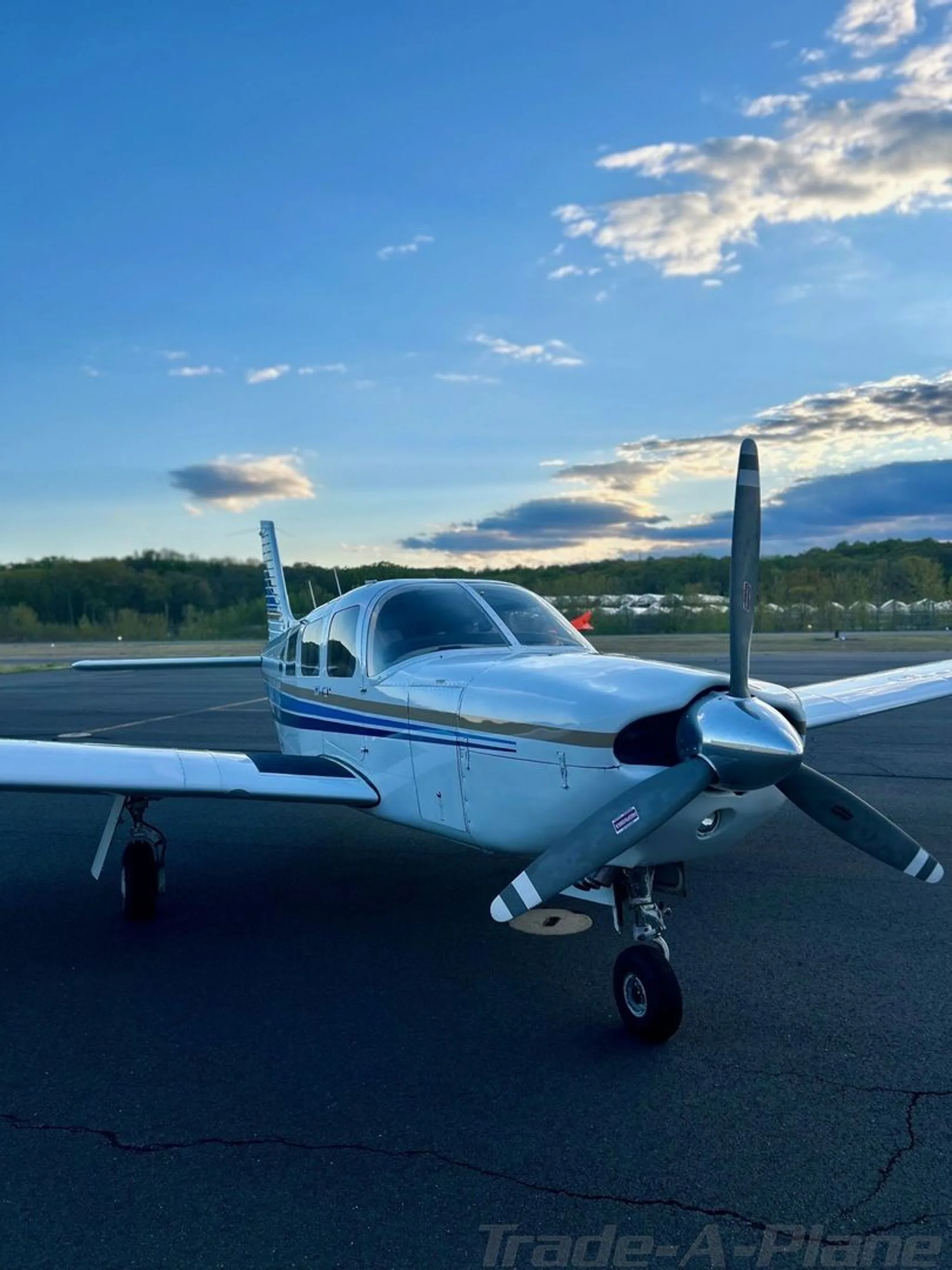 A front three-quarter view of a white Piper Cherokee aircraft with a propeller, parked on an airfield at sunset.