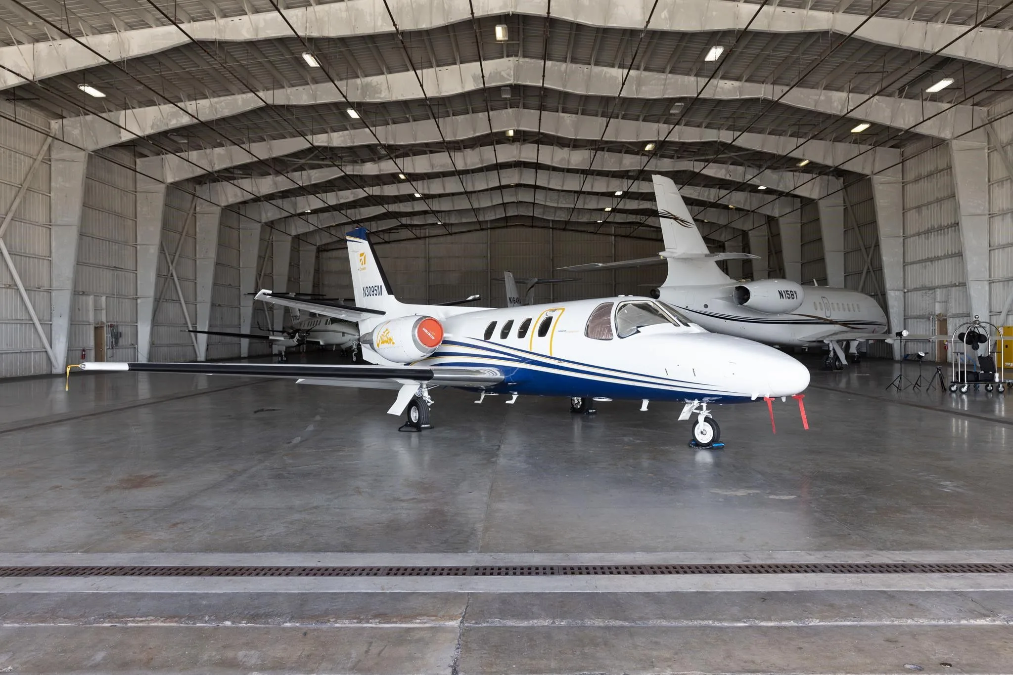 A white and blue Cessna Citation jet (N3095M) parked inside a large hangar with another jet in the background.