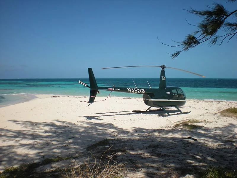A dark green Robinson R44 Clipper II helicopter, registration N4520X, is parked on a sunny white sand beach with turquoise water in the background.