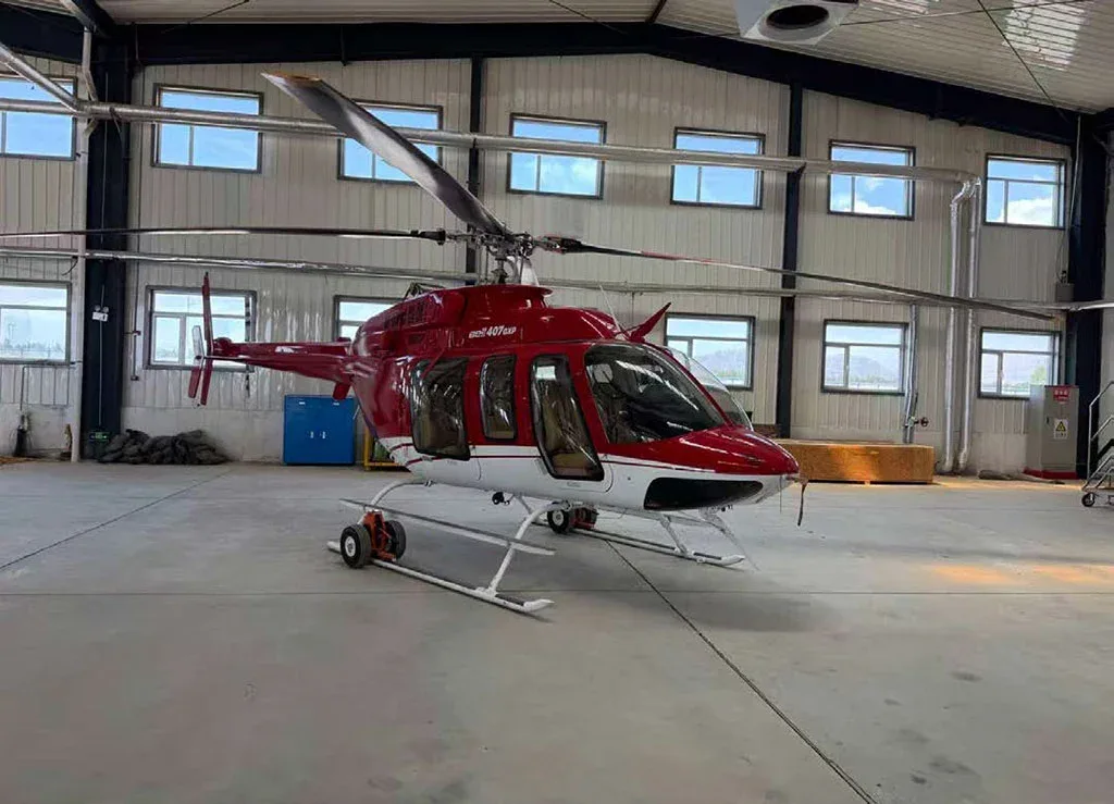 A red and white Bell 407GXP helicopter is parked inside a hangar with many windows.