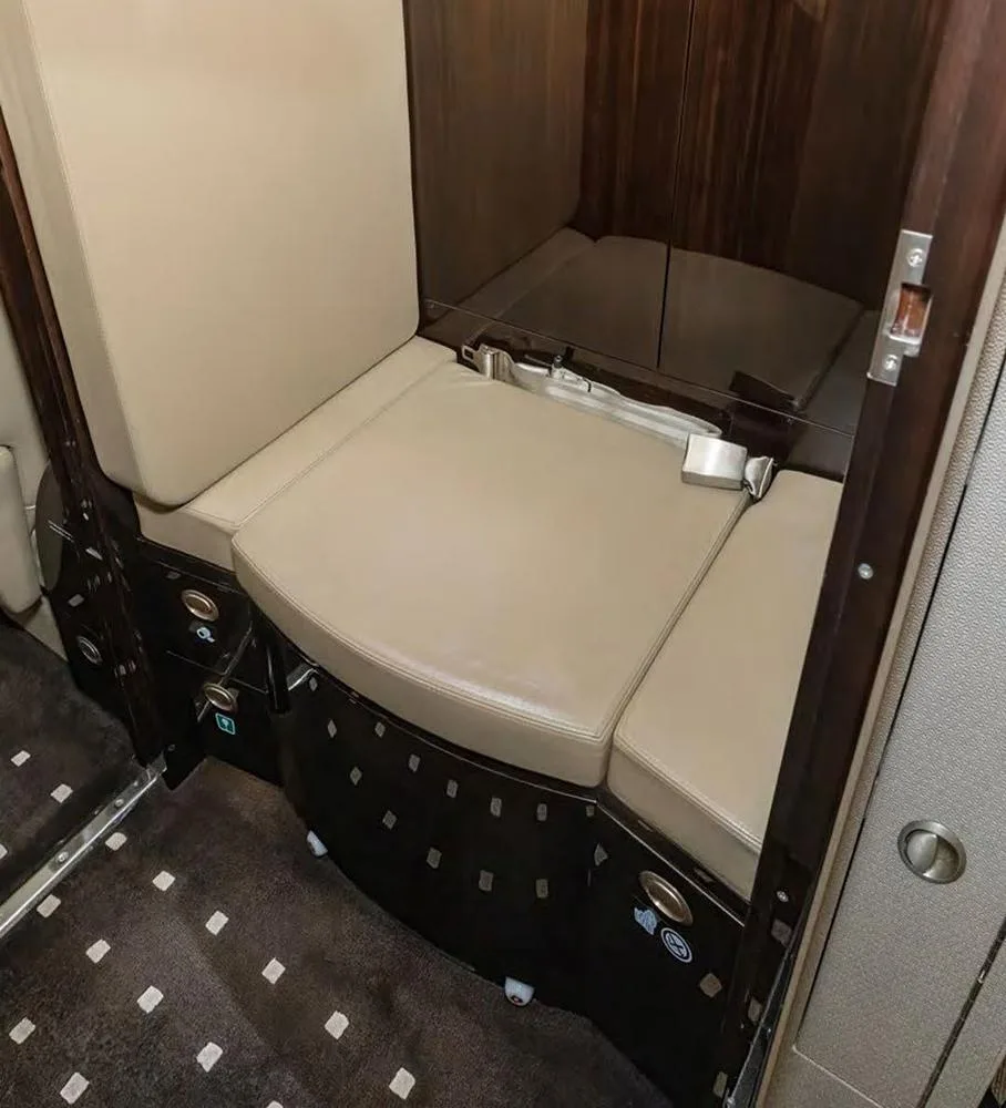 A small beige leather seat or divan with a seatbelt, set against dark wood paneling in the aircraft cabin.